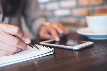 Closeup image of a woman's hands writing on notebook and using and pointing at smartphone on wooden table in modern cafe
