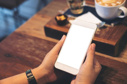 Mockup image of a woman's hands holding white mobile phone with blank screen with coffee cup on wooden table in vintage cafe