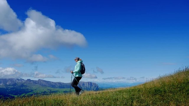 Woman-hiker descends the green slope agains a backdrop of mountains and blue sky