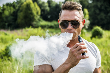 Handsome man standing in nature and vaping with electronic cigarette. 