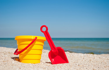 Fototapeta na wymiar Yellow childrens bucket with red toy toy plastic red scapula on the left against the blue sea and blue sky yellow sand beach sea shore seashells summer vacation sunny summer day, bright sun, baby toys