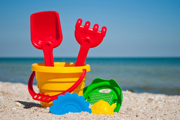 Yellow baby bucket with red handle, plastic red spatula and rake, green sieve, yellow sand form of strawberry, blue plate, blue sea and sky, yellow sand beach seashells seaside summer vacation sunny