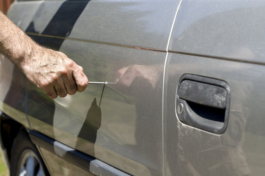 Ręka trzyma gwóźdź i niszczy lakier samochodu na drzwiach rysując krechę.
