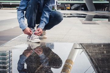 handsome man sit on concrete floor tying shoelace to wearing the shoes with  water reflection on floor. - obrazy, fototapety, plakaty