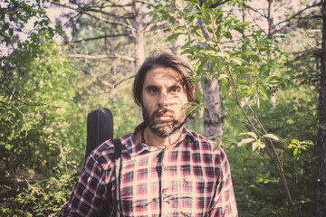 Naklejka premium handsome male with beard and long hair walking in the summer forest with guitar case 