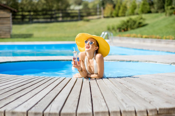 Young woman in swimsuit with big yellow sunhat relaxing with a bottle of fresh drink sitting on the poolside outdoors