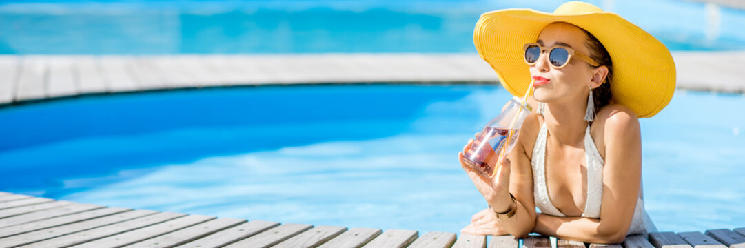 Young woman in swimsuit with big yellow sunhat relaxing with a bottle of fresh drink sitting on the poolside outdoors. Panoramic cropped image