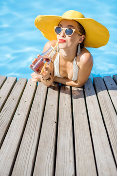 Young woman in swimsuit with big yellow sunhat relaxing with a bottle of fresh drink sitting on the poolside outdoors
