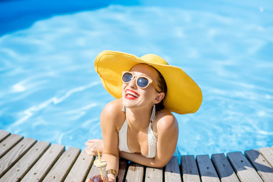 Young woman in swimsuit with big yellow sunhat relaxing with a bottle of fresh drink sitting on the poolside outdoors