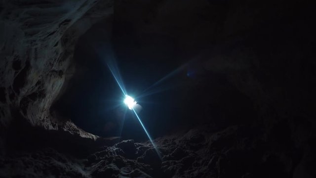 Young man with hard effort stretches out his hand trying take flashlight which lies on the ground in cave. Absolutely deep darkness around. Traveler inside the cave.