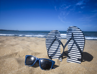 Blue sandal flip flop and sunglasses on the sand beach with blue sea and sky background in summer...