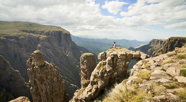 A male hiker sitting on rock stage with a natural bridge above an epic mountainous valley.