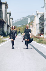 Two Stylish fashionable girl with blonde long hair,  stylish black dress and modern handbag walking in the center of the street. Fashion trendy woman concept