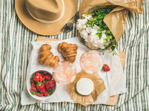 French style summer outdoor picnic setting. Flat-lay of glasses of rose wine, strawberries, croissants, brie cheese on board, straw hat, sunglasses, peony flowers, top view. Outdoor gathering concept