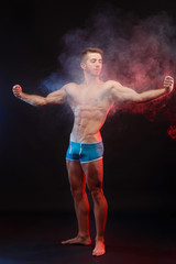 Young handsome muscular man bodybuilder with perfect abs, shoulders,biceps and triceps demonstrating huge muscles in the smoke, full length portrait