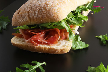 Ciabatta sandwich with jamon ham serrano paleta iberica, arugula, rosemary on stone slate black background