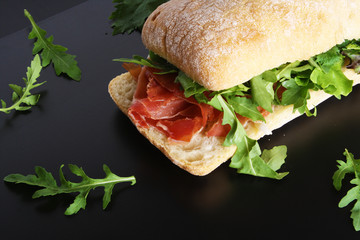Ciabatta sandwich with jamon ham serrano paleta iberica, arugula, rosemary on stone slate black background