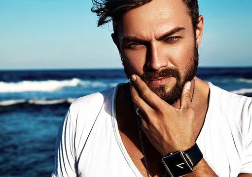 Portrait of handsome fashion man model wearing white clothes posing on blue sea background. Touching his beard
