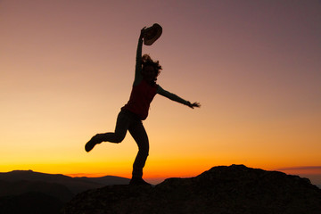 Silhouette of happy joyful young attractive woman jumping and having fun at the mountain against the sunset. Freedom, adventure and leisure vacation concept.