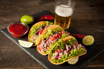 Photo of Mexican tacos with ground meat, beef, beans, onions and salsa on wooden background. Ketchup sauce and lime. A glass o beer in the background.