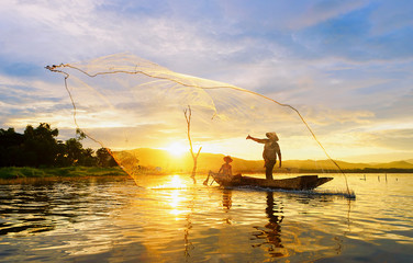 Fisherman on wooden boat casting a net for catching freshwater fish in reservoir.