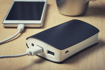 Closeup of a powerbank charging a smartphone on a wooden table next to a cup of coffee