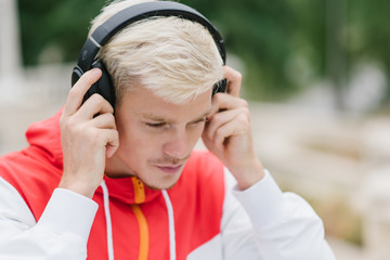 Shot of attractive runner with muscular athletic body dressed in red sportswear resting after physical activities in open air in park. Young handsome man listening to music with headphones outdoor.