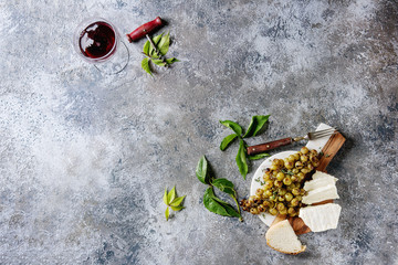 Obraz na płótnie Canvas Serving board with sliced camembert cheese and baked bunch of green grapes served with bread, glass of red wine, corkscrew, green leaves, fork over gray texture background. Top view with copy space