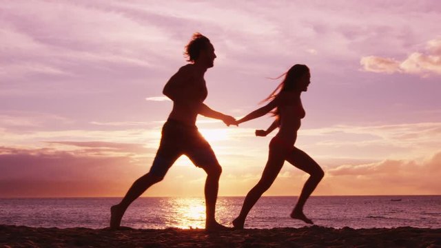 Beach couple running having fun holding hands on beach enjoying romantic sunset on summer travel vacation holiday honeymoon. Playful young woman and man in love. RED EPIC SLOW MOTION.