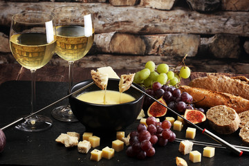 Gourmet Swiss fondue dinner on a winter evening with assorted cheeses on a board alongside a heated pot of cheese fondue with two forks dipping bread and white wine behind in a tavern or restaurant