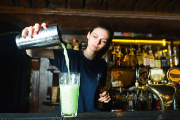 Barmen cakes a cocktail at the bar. A bartender girl pours a shaker ingredient into a glass of cocktail.