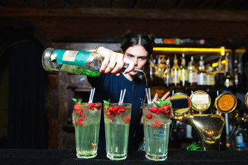 A bartender girl creates beautiful alcoholic cocktails with berries in a nightclub. The barman drinks bottled alcohol in a cocktail.
