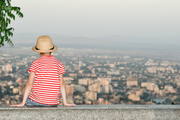 Boy sitting and looking at the city from a height. Rear view, evening time