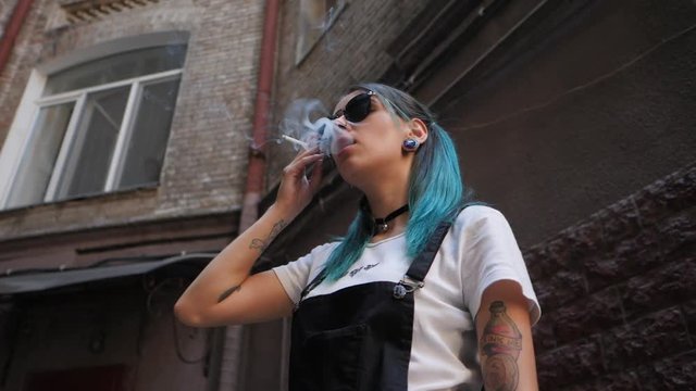 SLOW MOTION: Homeless emo girl smoking cigarette.Street punk or hipster in sunglasses with blue dyed hair,piercing,lenses,tattooed,ears tunnels and unusual hairstyle stands in backyard.