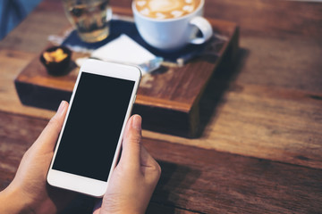 Mockup image of a woman's hands holding white mobile phone with blank black screen with coffee cup , tea and snack on wooden table in vintage cafe