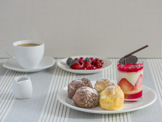 A dish of chou cream and a glass of strawberry chease cake with a cup of tea or coffee and a dish of various berry for an afternoon break.