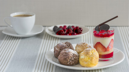 A dish of chou cream and a glass of strawberry chease cake with a cup of tea or coffee and a dish of various berry for an afternoon break.