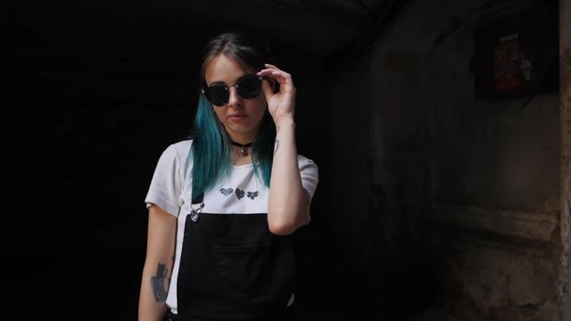 SLOW MOTION: Homeless emo girl posing and corrects sunglasses.Street punk or hipster with blue dyed hair,piercing,lenses,tattooed,ears tunnels and unusual hairstyle stands in backyard.