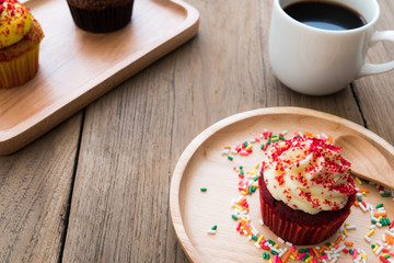 Red cupcakes put on a spherical wooden plate. Beside of cupcake have white coffee mug.In the background have yellow cupcake and chocolate cupcake.All of it rests on wooden table.