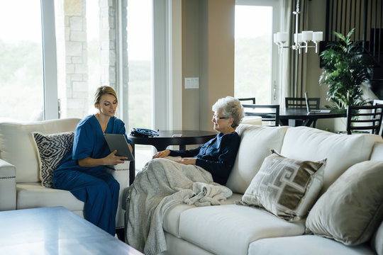 Home caregiver showing tablet computer to senior woman while sitting on sofa in living room