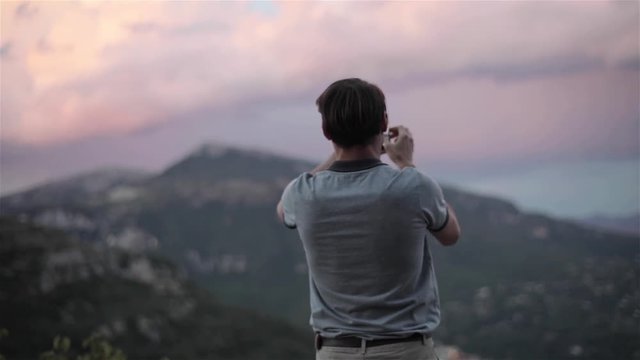Male traveller takes photo with phone of pink clouds sky in mountains slow motion close up from back and side rack focus, looking for pictures touches screen focuses enjoys view and app possibilities