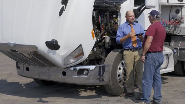 Insurance agent, or other official with electronic notepad, talks to truck driver during an inspection of truck engine. Full length view from front angle. Hand-held 4K