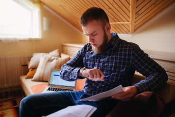 Young man studying from home sitting on a sofa holding documents while working on a laptop computer