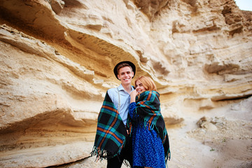 An attractive man hugs a beautiful woman. The pair wrapped in plaid in the middle of a sandy canyon