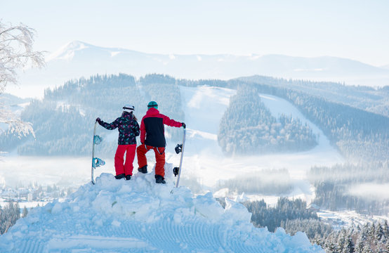 Two snowboarders enjoy the snow-white scenery of mountains and forests of the Carpathians from the height of the mountain top on a beautiful sunny winter day. Back view