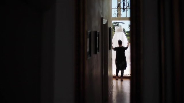 Woman stands in doorway checking dress hanging on terrace. Female silhouette of maid or dressmaker designer in light seen from dark corridor in old house