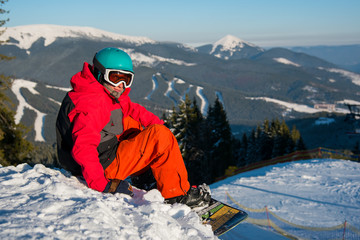 Fototapeta na wymiar Shot of a male snowboarder resting on top of the mountain, relaxing on the edge of a slope looking to the camera. Beautiful view of snowy mountains on the background. copyspace recreation tourism