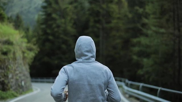 Man jogging on mountain road no face back close up slow motion. Alone runner outdoors in morning in grey hooded sweater. Running sport man sprints exercising and training in nature. Freedom and choice