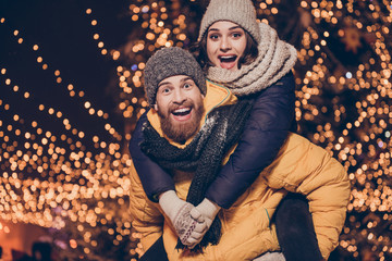 Love forever. Funky mood. Young handsome red bearded boyfriend is piggy backing his cute lover, wearing winter warm outfits, behind them are christmas illuminations