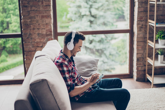 Side profile shot of cheerful hipster enjoying listening to his favourite song at his holiday in big white modern earphones, wearing checkered shirt, sitting on beige couch, big windows, loft interior